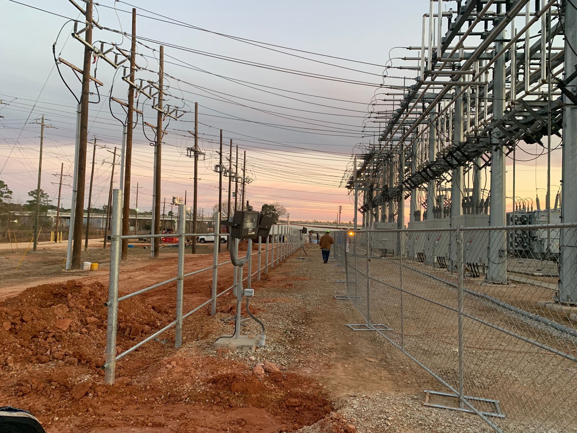 Substation construction site at sunset, with metal frameworks, poles, fencing, and workers in the distance