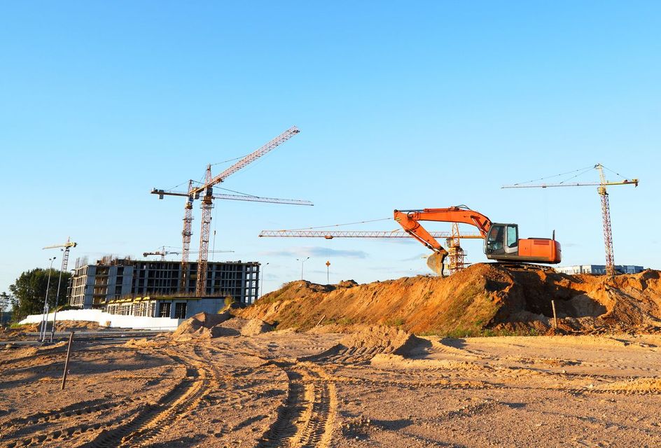 Construction site with cranes and an excavator on a dirt mound under a clear blue sky