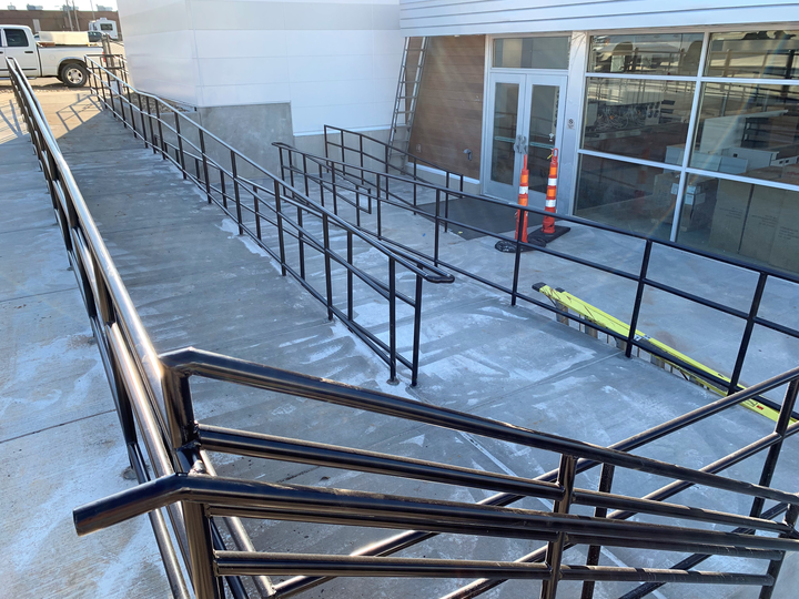 Accessible ramp and stairs outside a glass-front building, with black handrails and orange cones near the entrance.