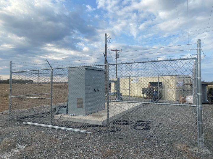 Chain-link fenced utility equipment pad with gray cabinet and electrical gear on gravel under cloudy sky