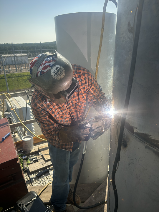 Worker in a hard hat welding near large tanks outdoors, with bright sparks and protective gear.