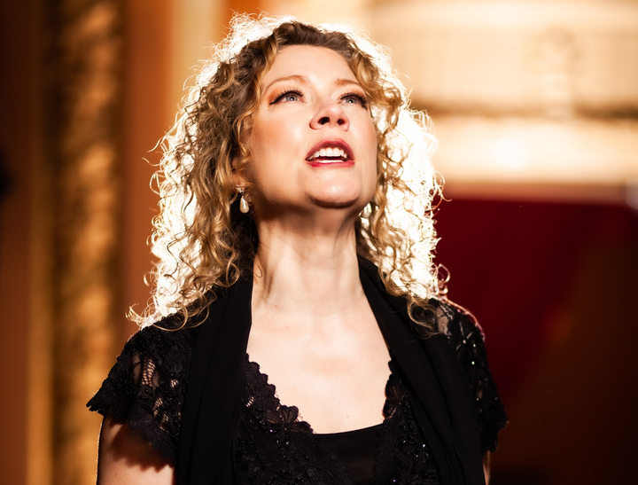 A woman with curly hair is singing into a microphone in a dark room.