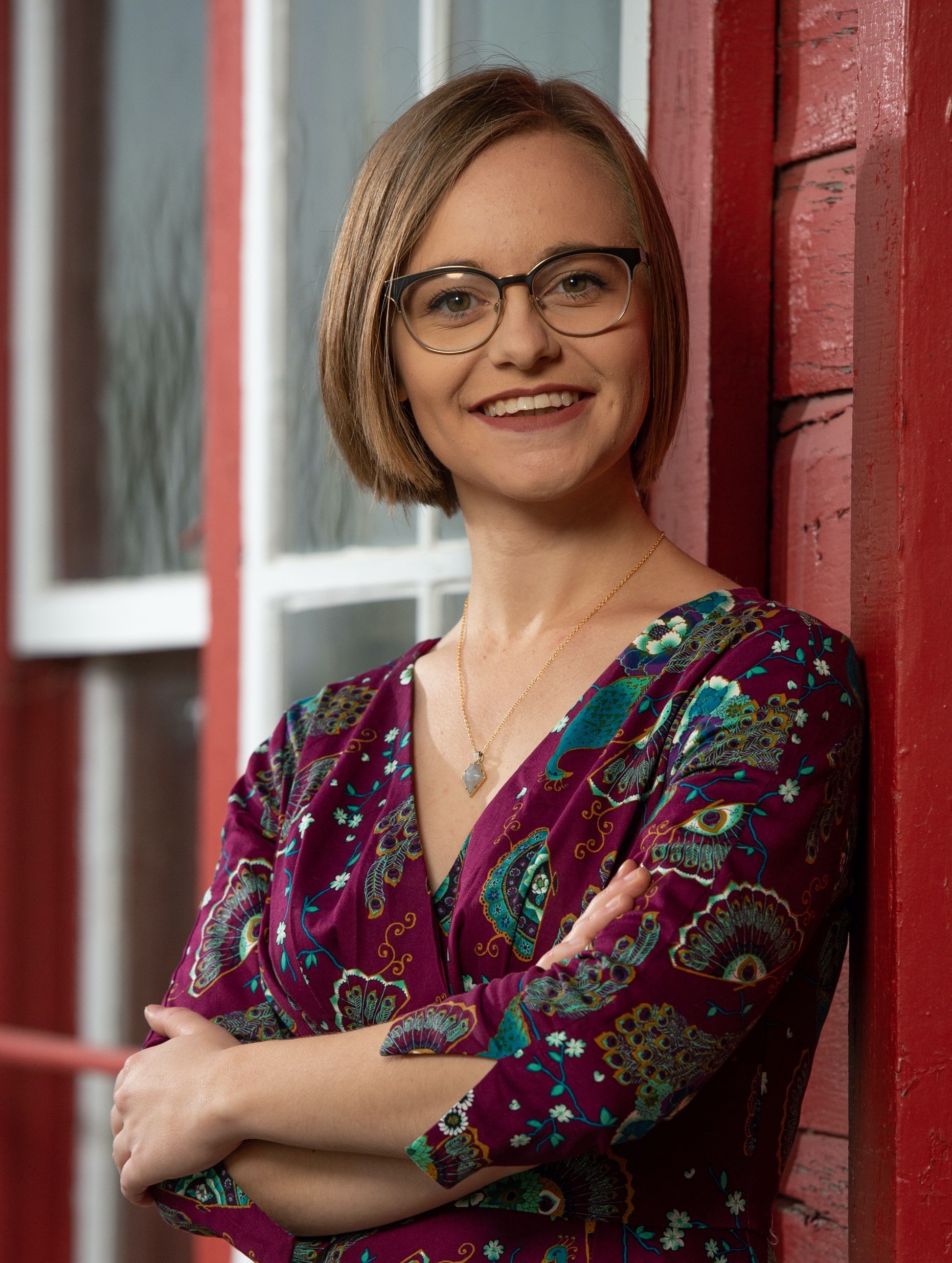 A woman wearing glasses and a purple dress is leaning against a red wall.