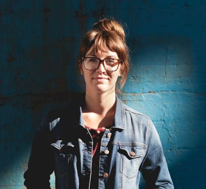A woman wearing glasses and a denim jacket stands in front of a blue wall