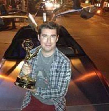 A man is sitting on the hood of a car holding a trophy.