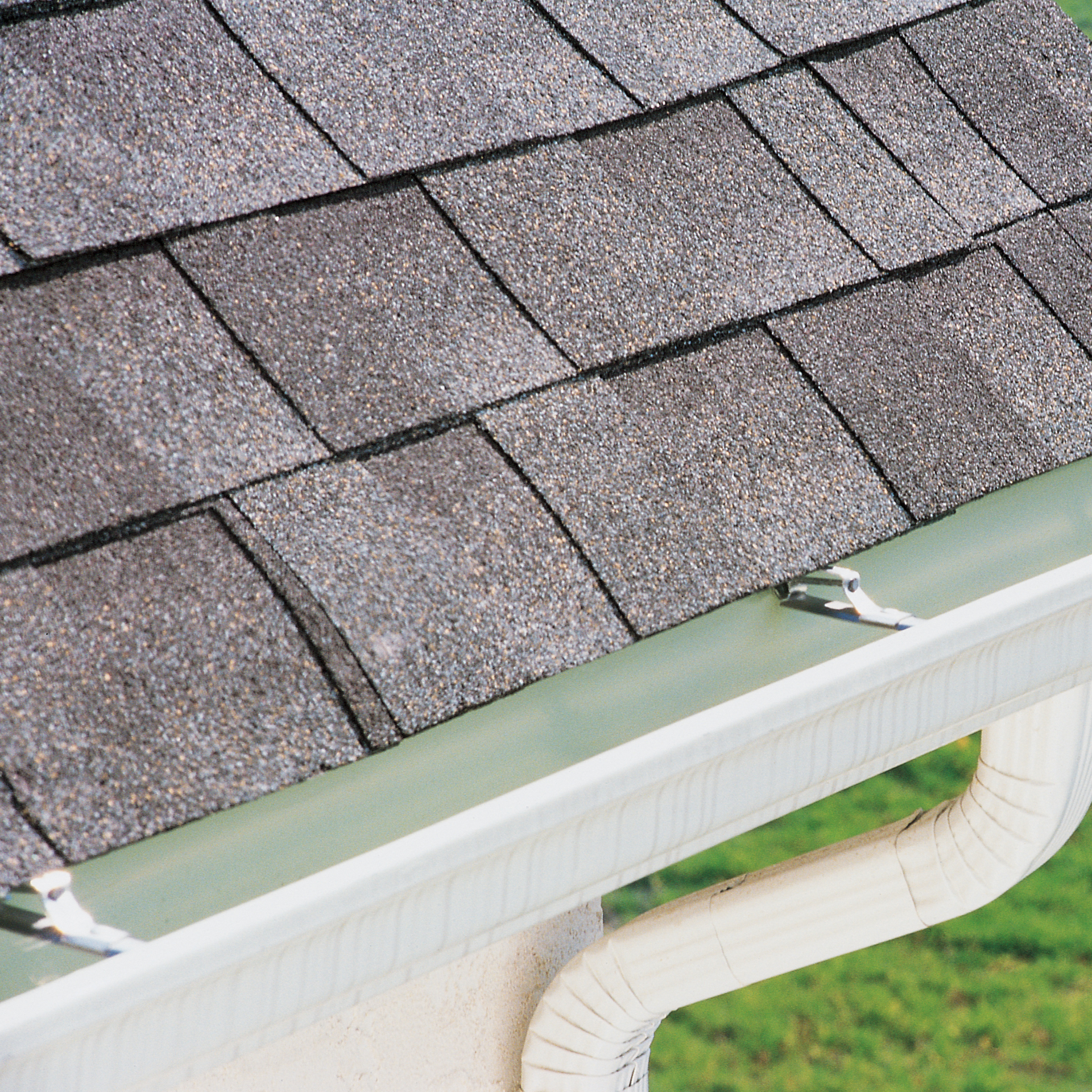 Gray asphalt shingles on a roof edge with white gutters and a matching downspout against a blurred green background.