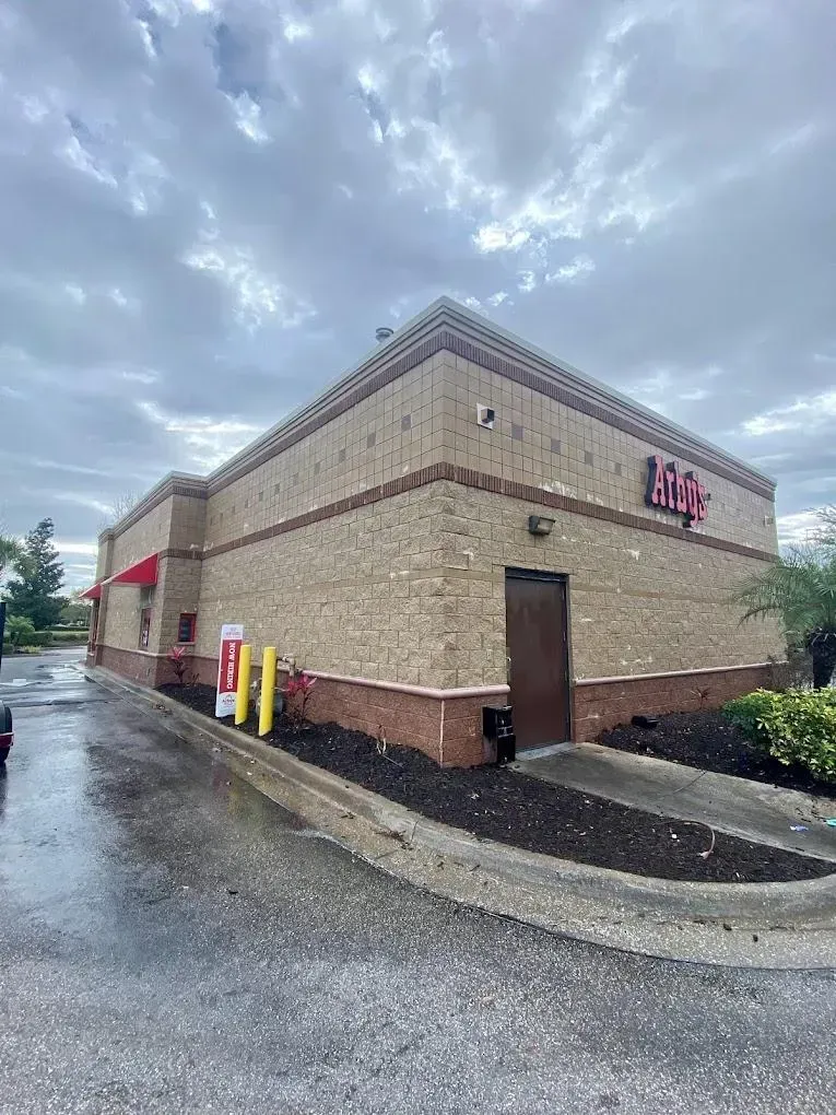 Exterior of a brick Arby's restaurant on a cloudy, rainy day with a drive-thru lane visible.