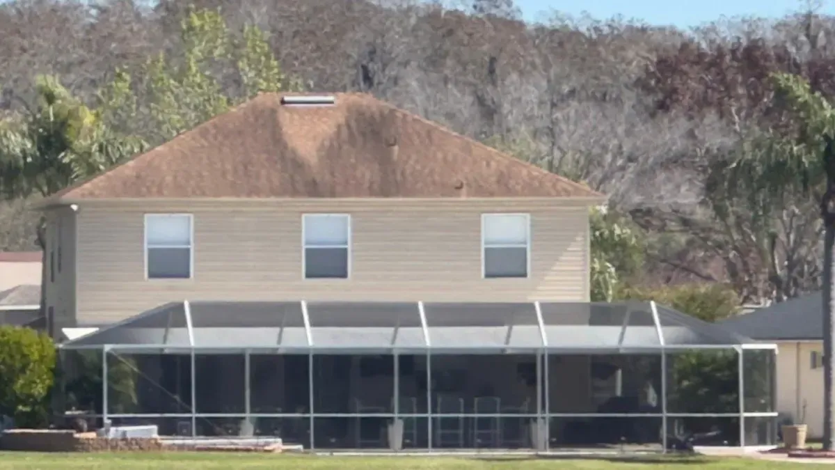A two-story tan house with a brown shingled roof and a large, screened-in porch, set against a backdrop of trees.