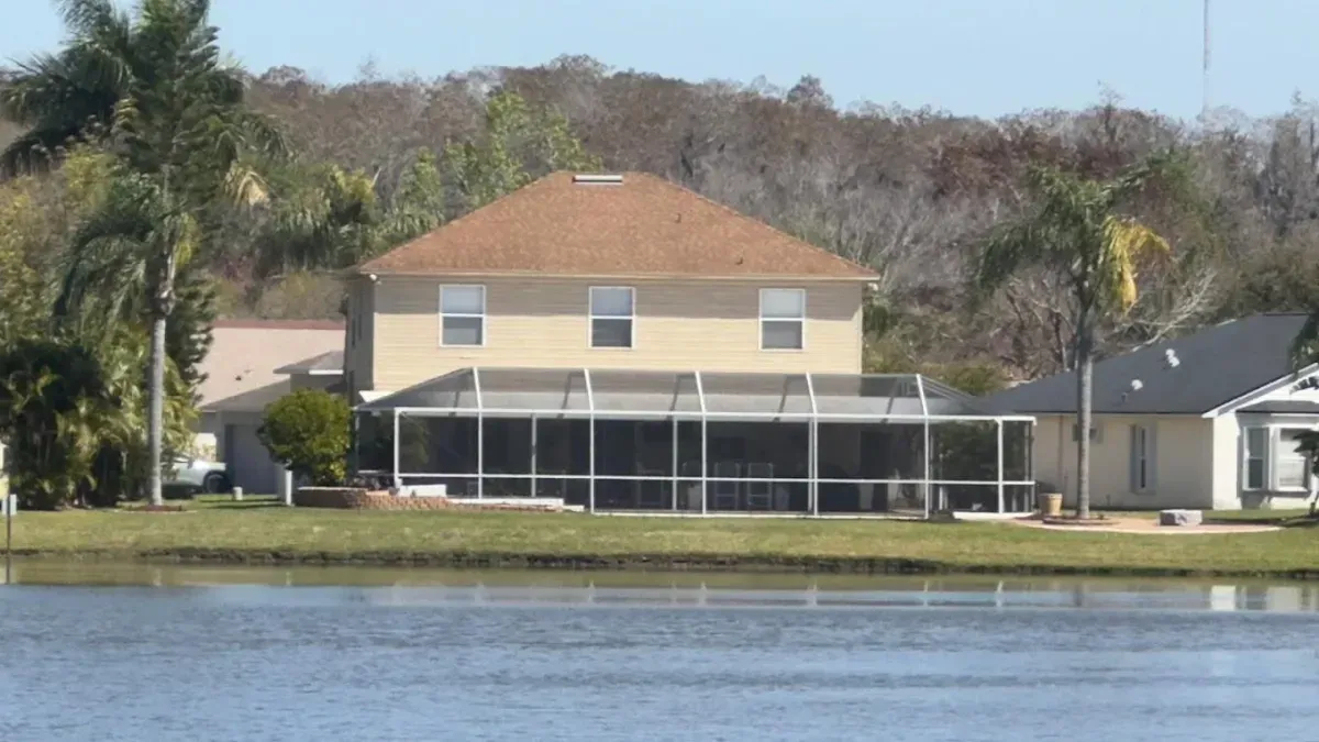 A two-story, tan-colored house with a brown roof and a large screened-in pool enclosure sits beside a calm lake.