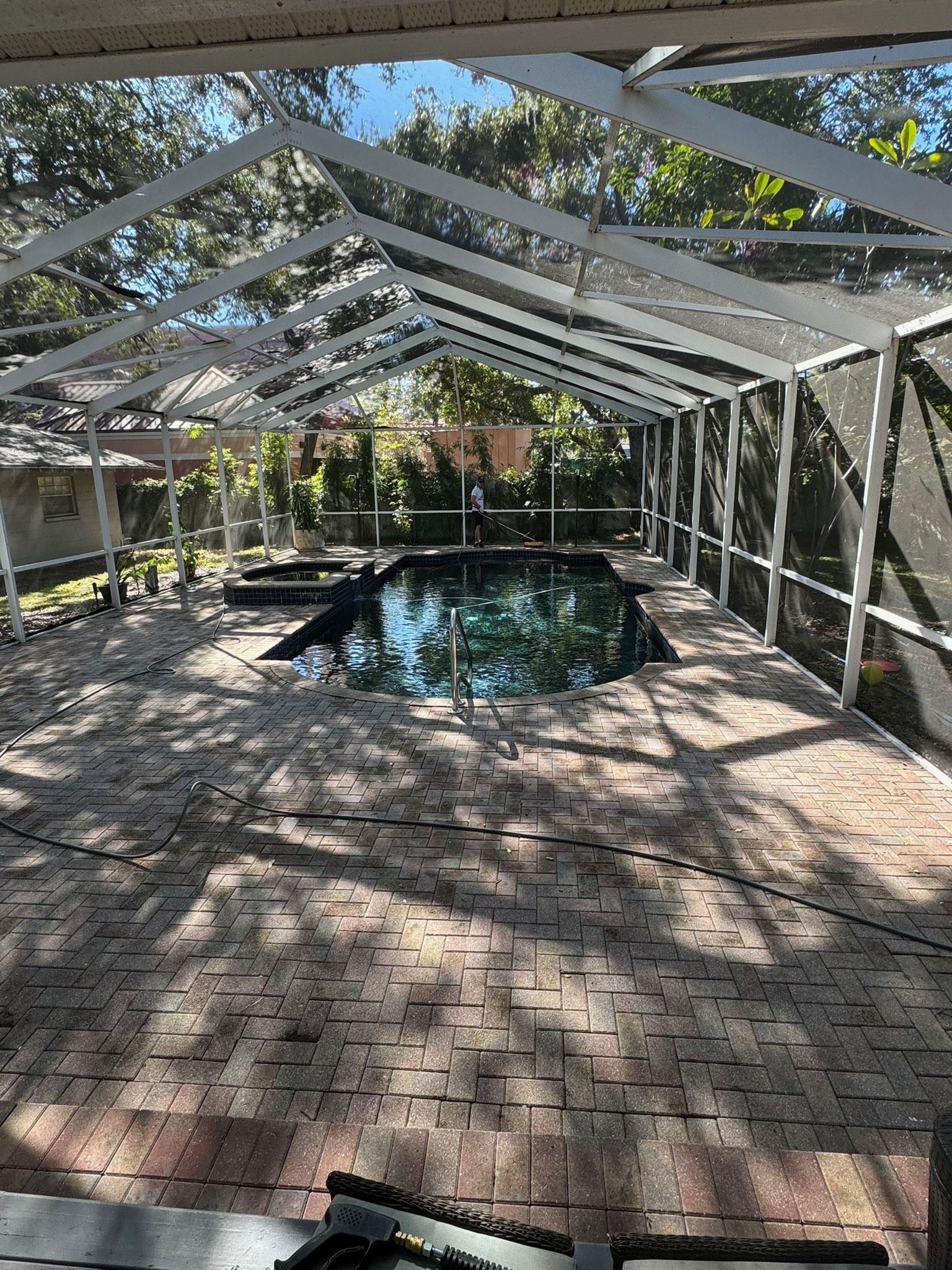 A backyard pool enclosed by a white-framed screen cage, featuring paved stone decking and surrounding trees.