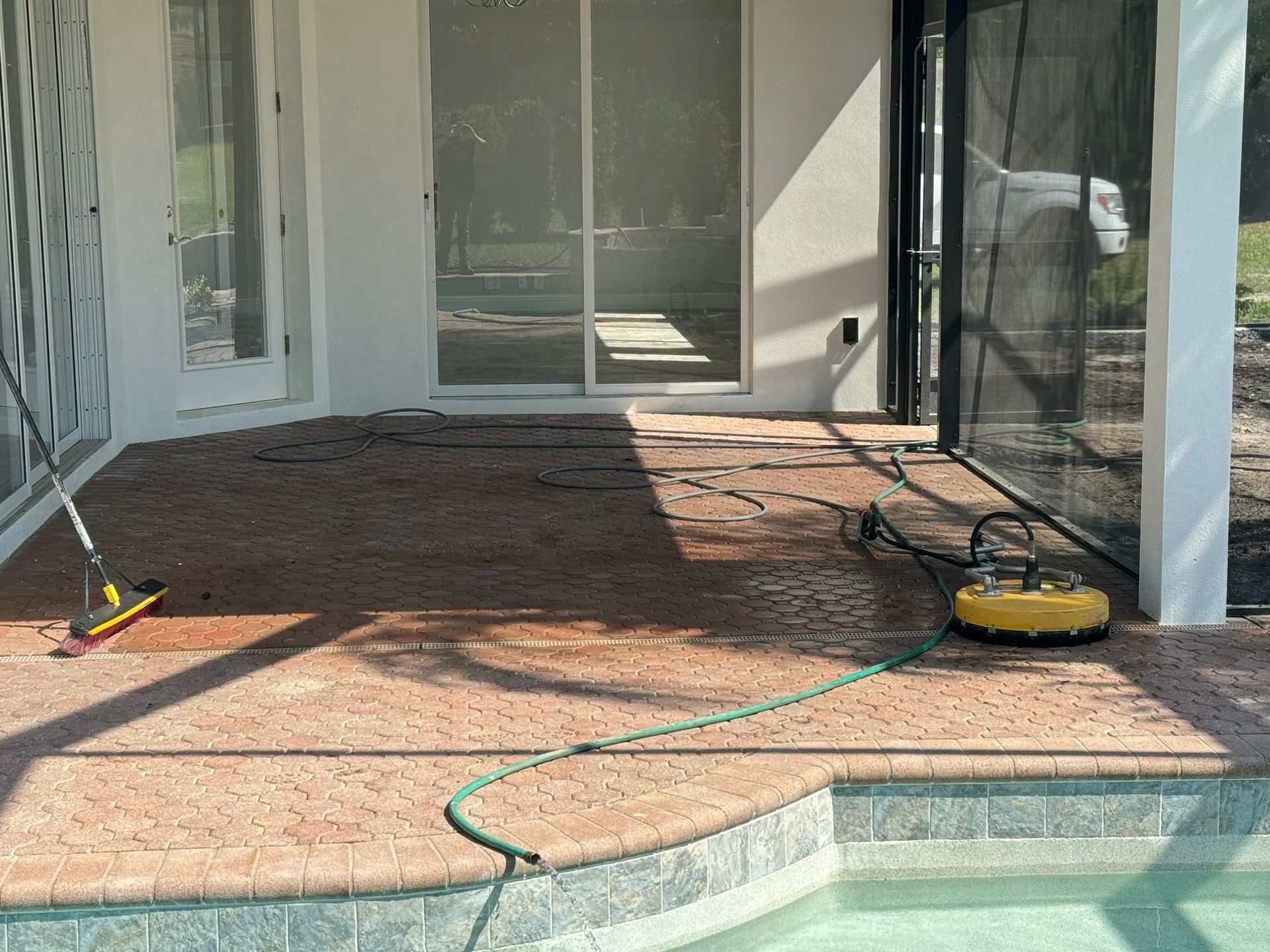 A patio floor with a pool deck being pressure-washed next to a sliding glass door and screened-in enclosure.
