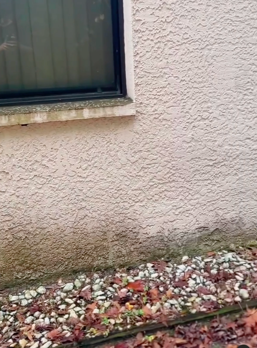 A close-up of a stucco wall showing water staining near the ground, with a window corner above and gravel at the base.
