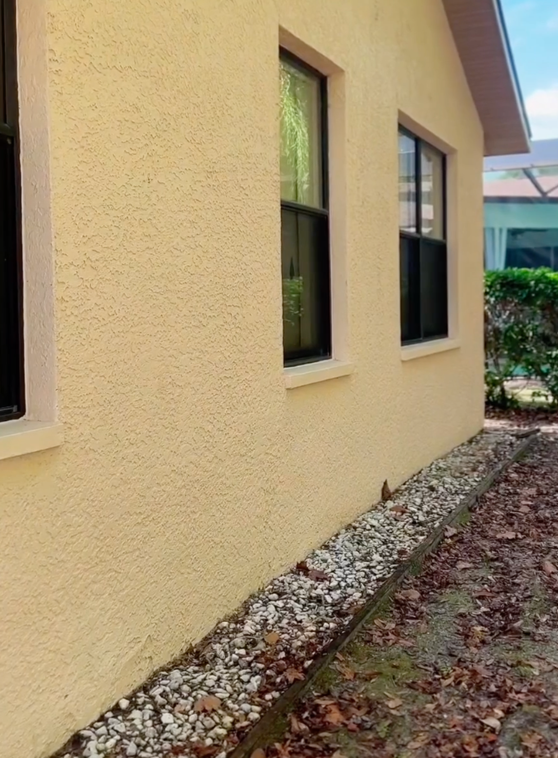 A beige stucco wall of a house with two dark-framed windows above a gravel garden bed covered in fallen leaves.