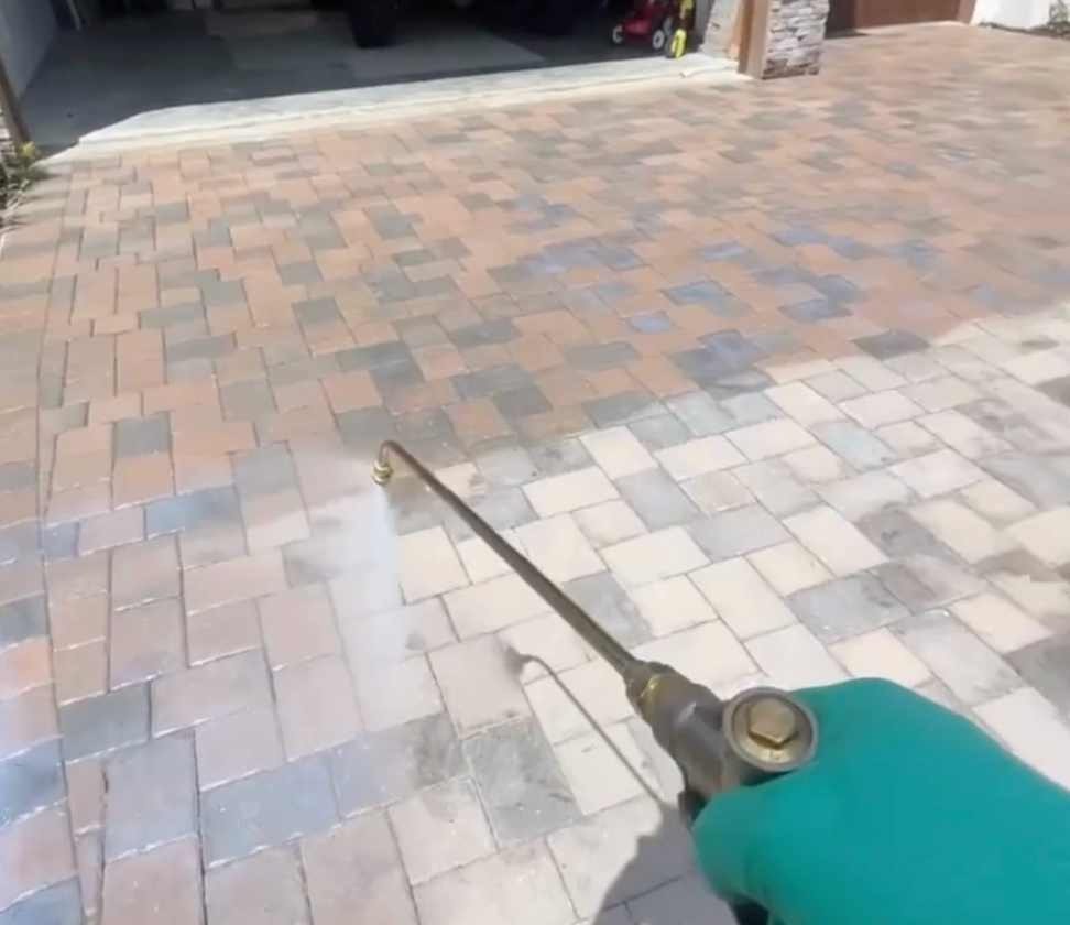 A gloved hand uses a pressure washer wand to clean a paved driveway, revealing a clear difference between dirty and clean.