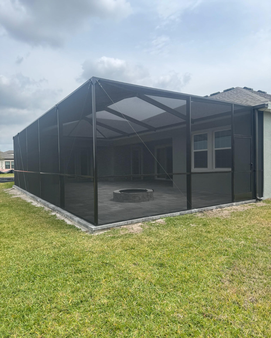 A black screen enclosure over a concrete patio featuring a small fire pit, viewed from the side in a residential backyard.