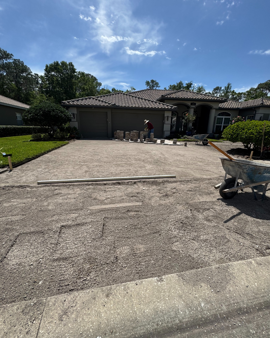 A worker installing pavers on a residential driveway on a sunny day, with a wheelbarrow in the foreground.