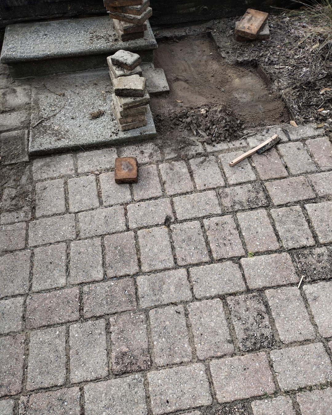A ground-level view of a stone patio showing a small area of removed pavers, a stack of bricks, and a hammer.