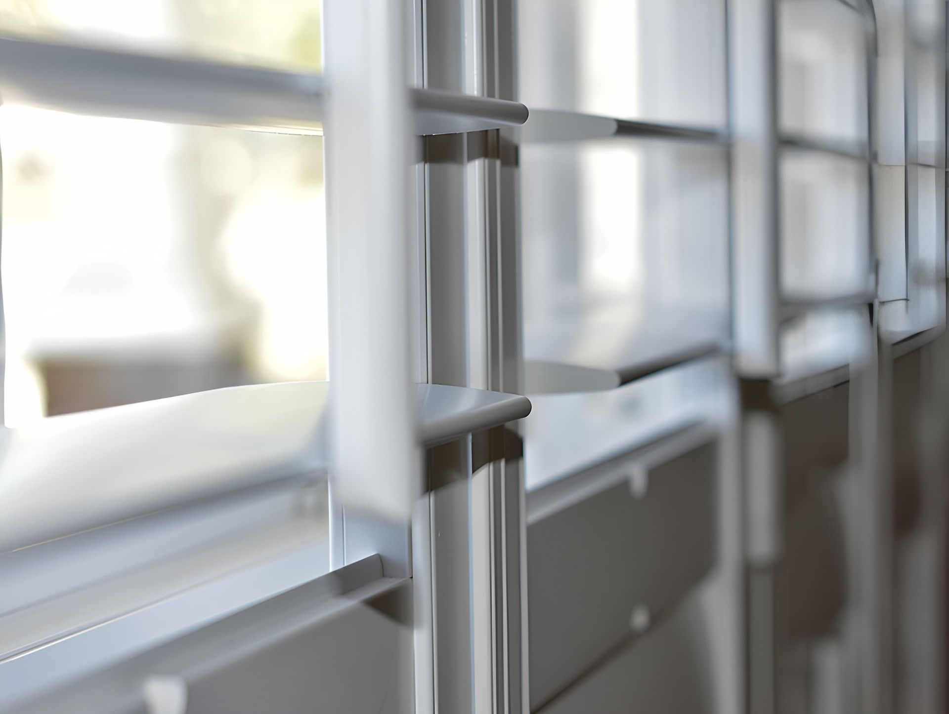 White window shutters, partially open, with a blurred background of a room and light — Manning Blinds & Awnings in Taree, NSW