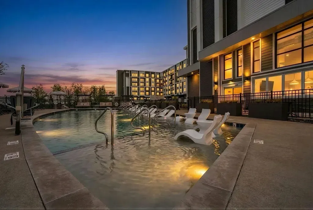 Modern apartment complex pool area with submerged lounge chairs and ambient lighting at dusk.