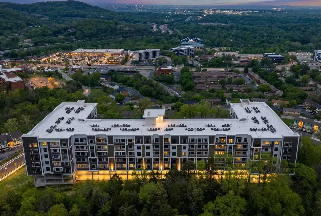 Aerial view of a modern apartment building exterior at dusk, with surrounding trees and distant city lights.