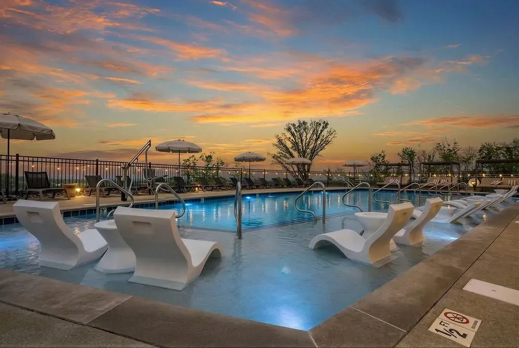 Outdoor pool deck with white lounge chairs and umbrellas at sunset, blue-lit water and colorful sky.
