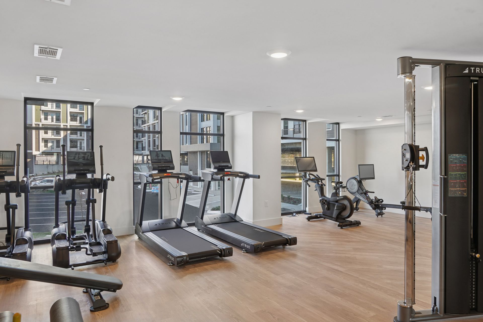 Bright gym with treadmills, weight machines, and wooden floors near large windows.