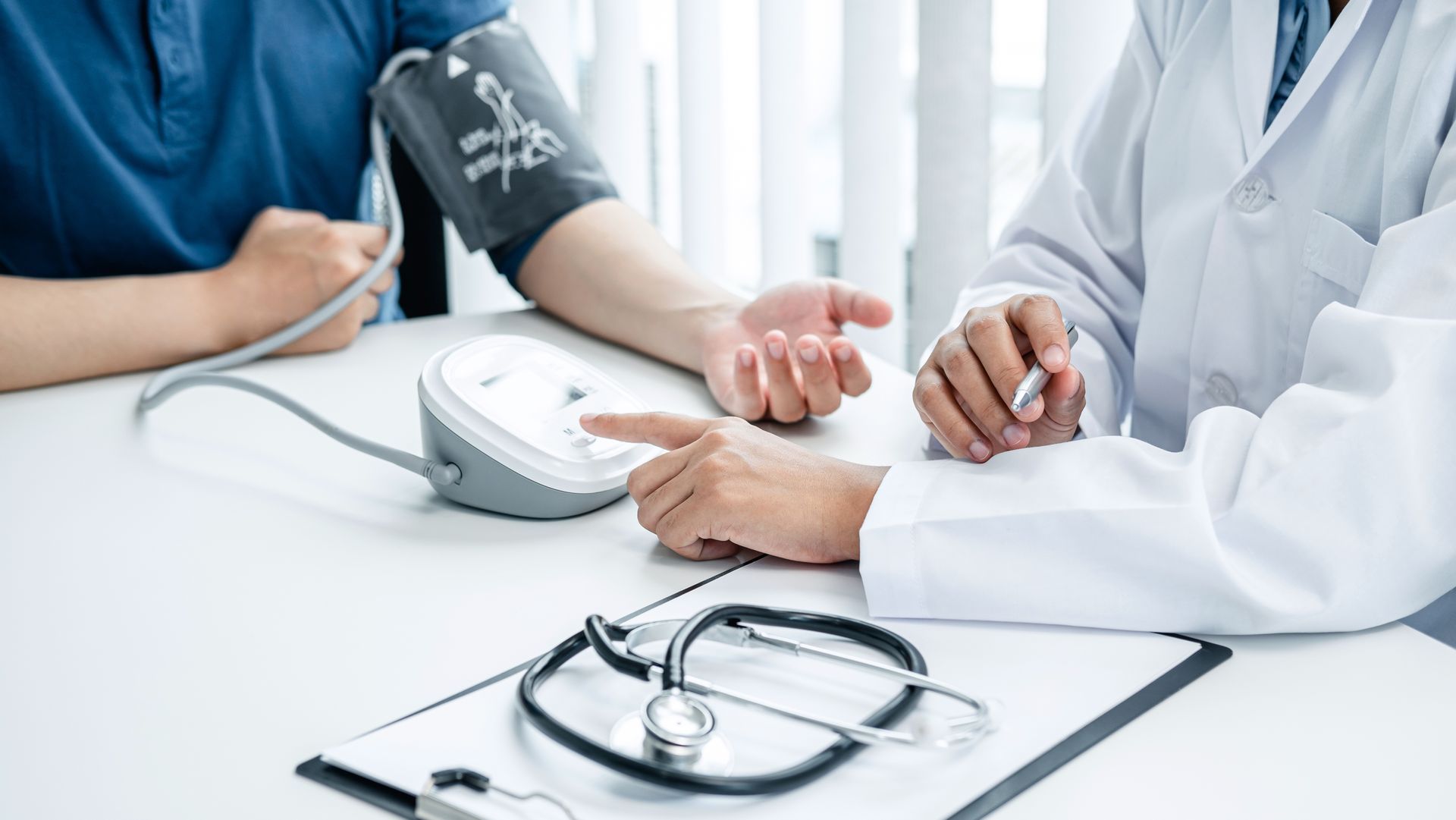 Person having blood pressure checked at a clinic desk with monitor and stethoscope visible.