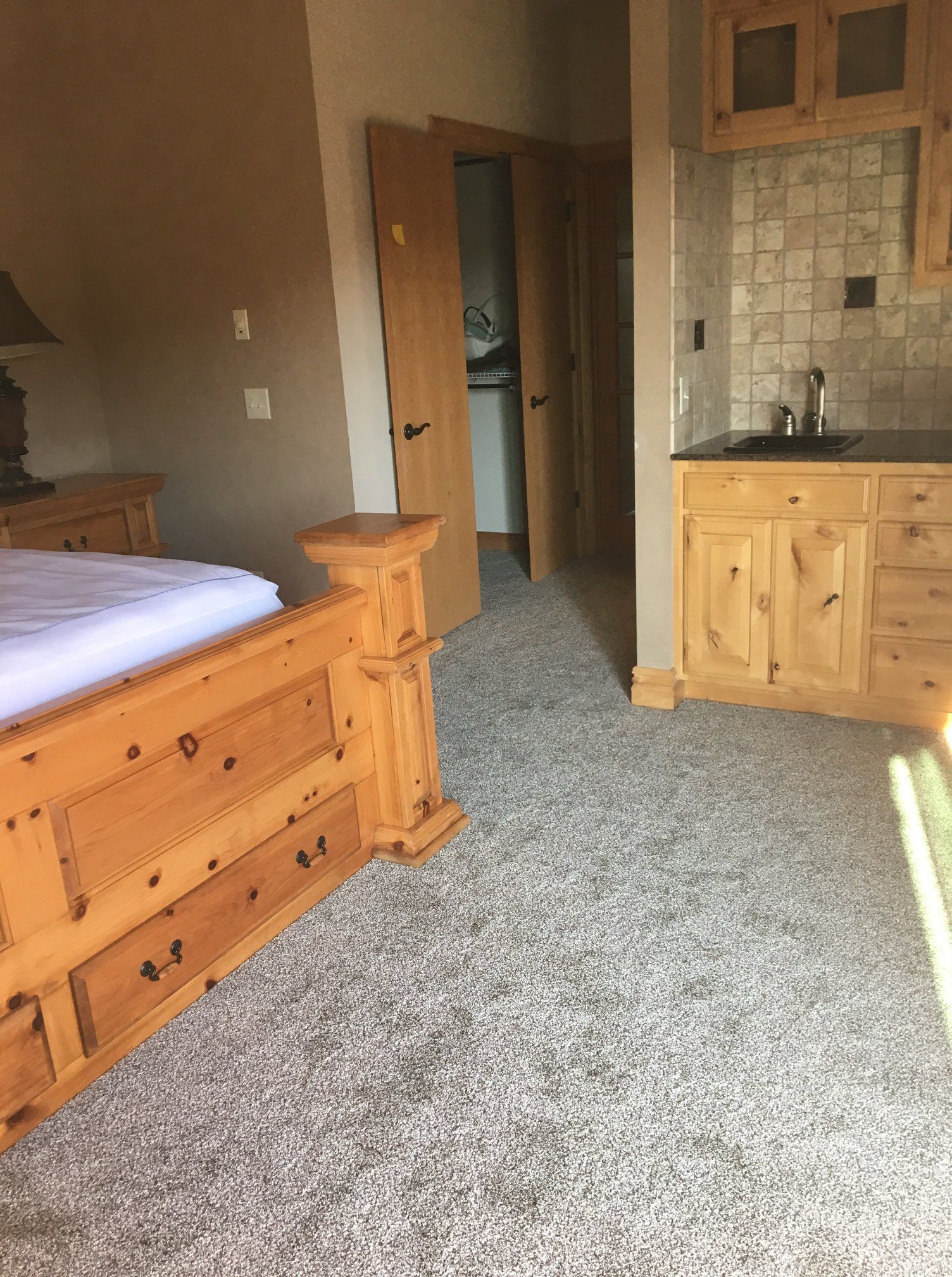 A room with a wood-framed bed, a small kitchenette with wood cabinets, and an open doorway leading to another room.