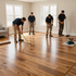 Four workers in navy shirts and khaki pants install wooden flooring in a brightly lit, empty room with two windows.