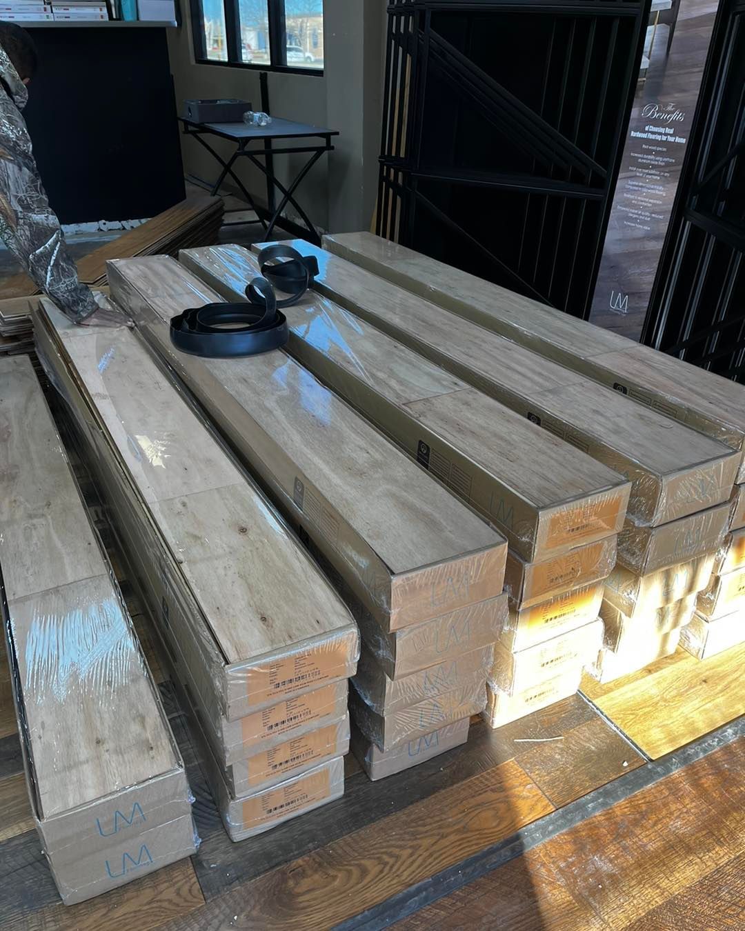 Stacked bundles of light wood flooring planks wrapped in clear plastic on a dark wooden floor.