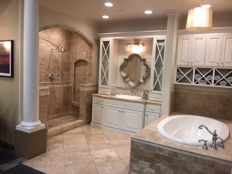 A luxurious bathroom featuring tan stone tiles, a walk-in shower with an arch, a white vanity, and a large soaking tub.