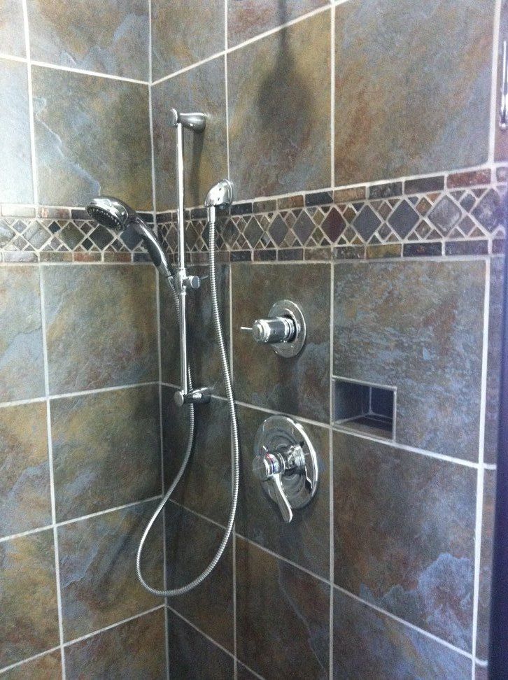 A shower stall with multicolored slate-style tiles, a chrome handheld shower head on a slide bar, and metal controls.
