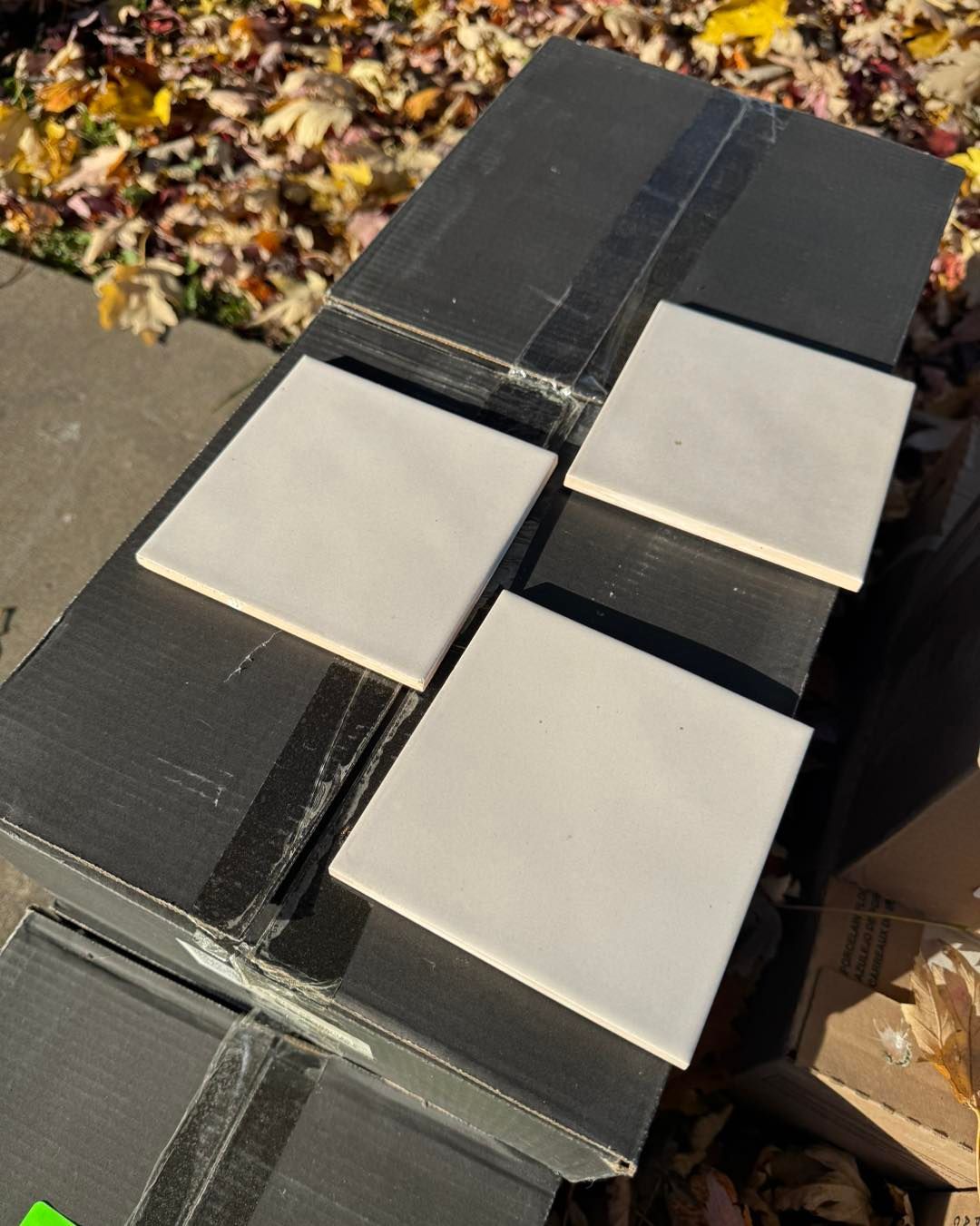 Three square, light-colored ceramic tiles sit on top of several dark shipping boxes outdoors near fallen leaves.
