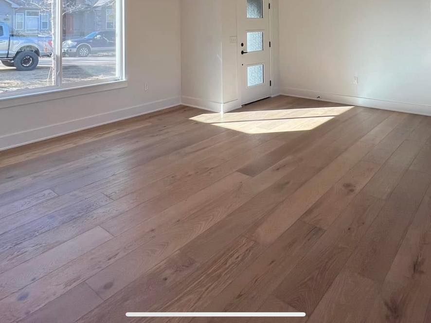A bright, empty room with light wood flooring, a window on the left, and a white interior door on the back wall.
