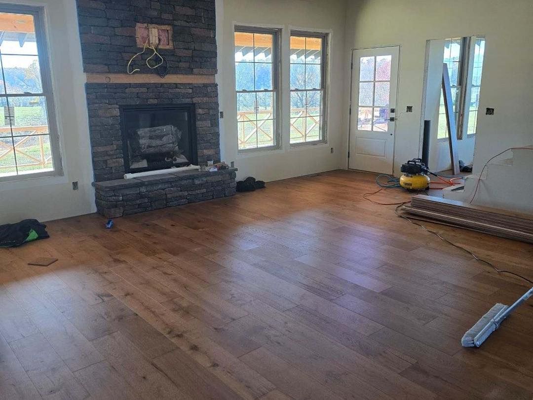 A newly installed hardwood floor in a room featuring a stone fireplace, large windows, and a white door.