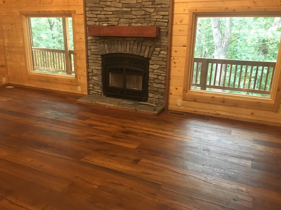 Rustic room with wide-plank wood flooring, a stone fireplace, and large windows overlooking a wooden deck and trees.