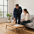 A professional in a blue uniform shows flooring samples to a person sitting on a sofa in a bright, modern living room.