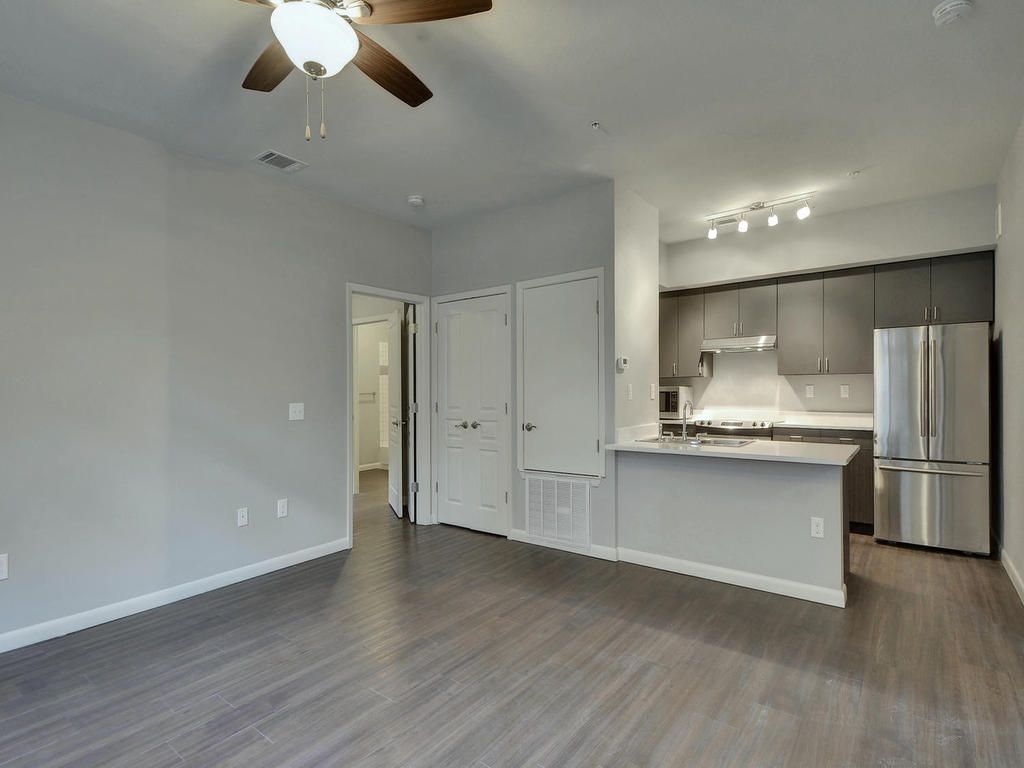 A living room with a ceiling fan and a kitchen in the background.