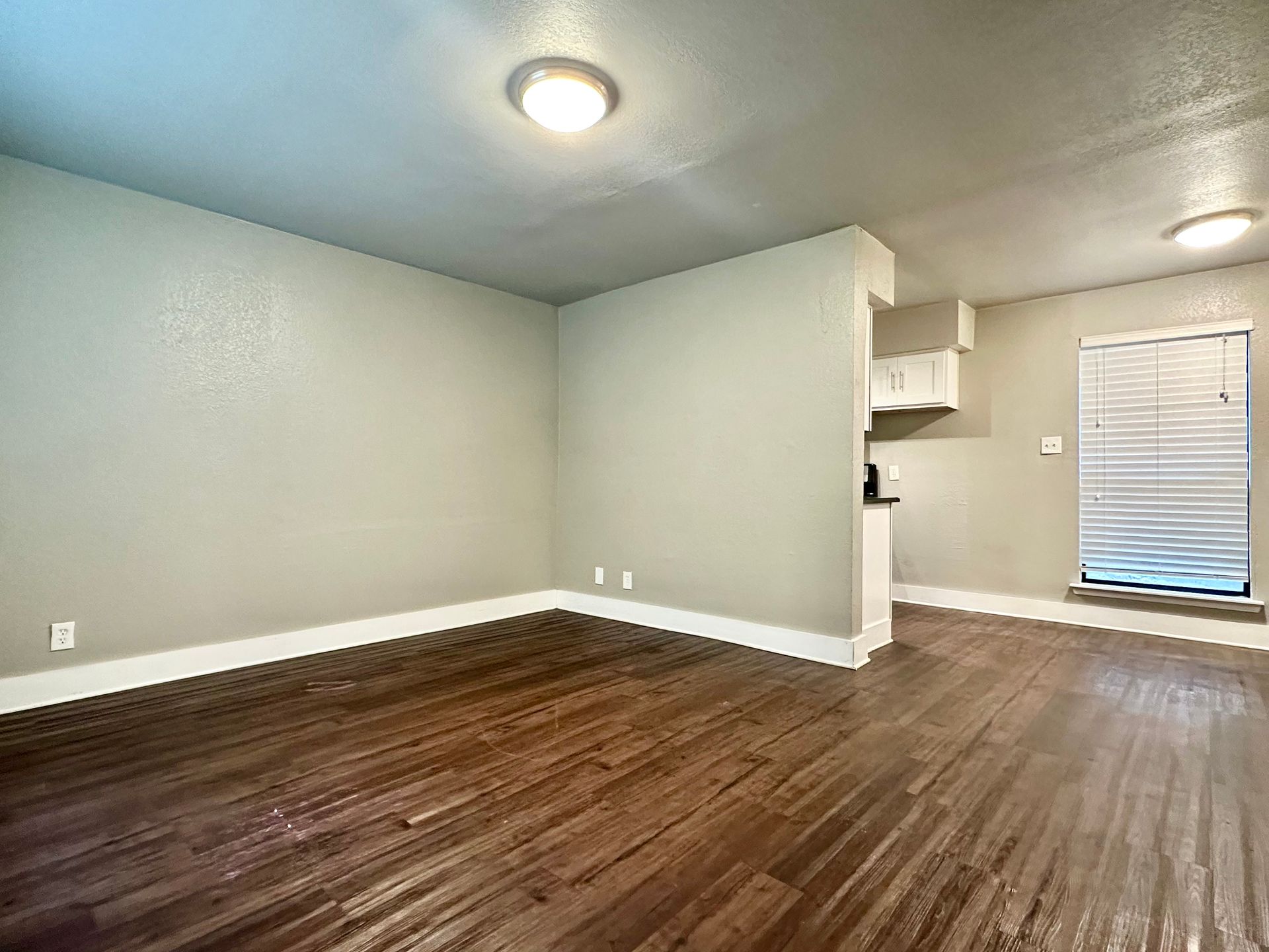 An empty living room with hardwood floors and a kitchen in the background.