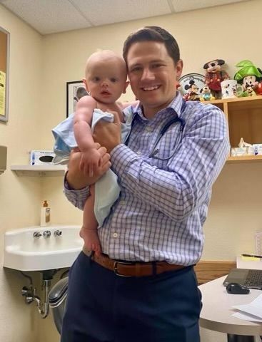Man holding a baby in a medical office, both facing the camera and smiling.