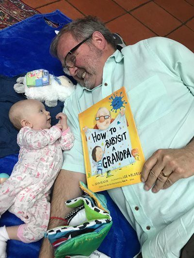 Man lying on a bed reads to a baby reaching toward the book, with a stuffed dinosaur nearby.