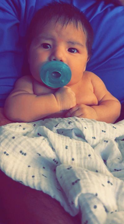 Baby lying on a blue blanket, holding a pacifier, with a white patterned blanket over the legs.