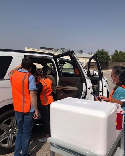 Two people in orange safety vests load supplies into a white SUV in a sunny outdoor setting