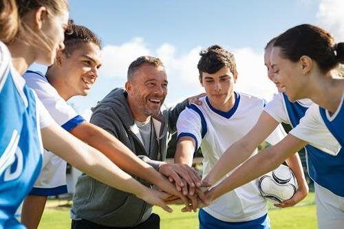 Soccer team huddling hands together on a sunny field with their coach, smiling before practice