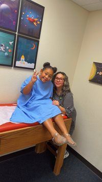 Two people sit on an exam table, smiling and flashing peace signs in a medical room.