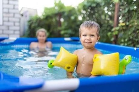 Child in blue pool with yellow floaties, smiling with an adult in the background near a brick wall.
