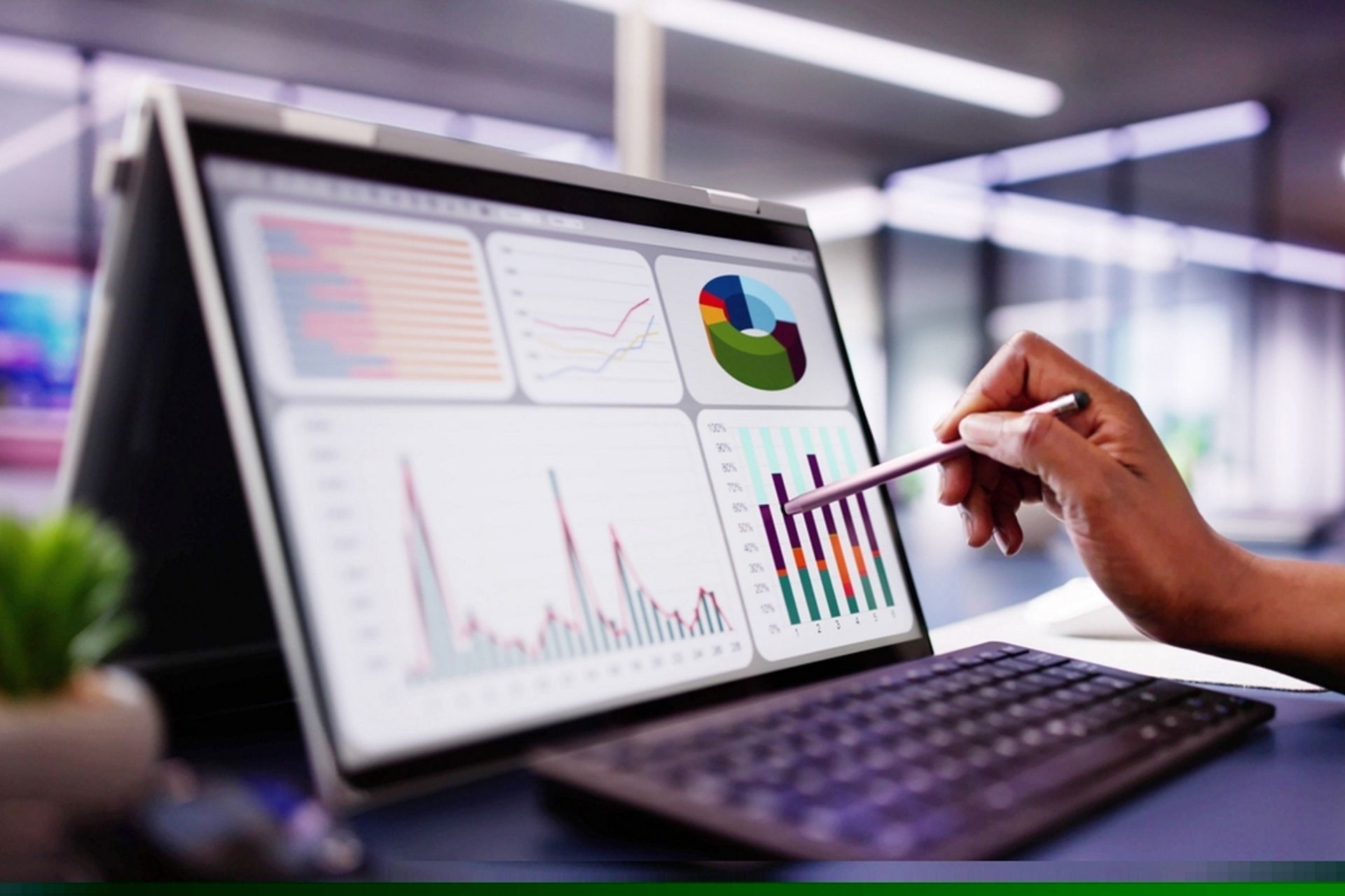 A hand uses a stylus to point at data visualizations on a digital tablet screen in an office setting.