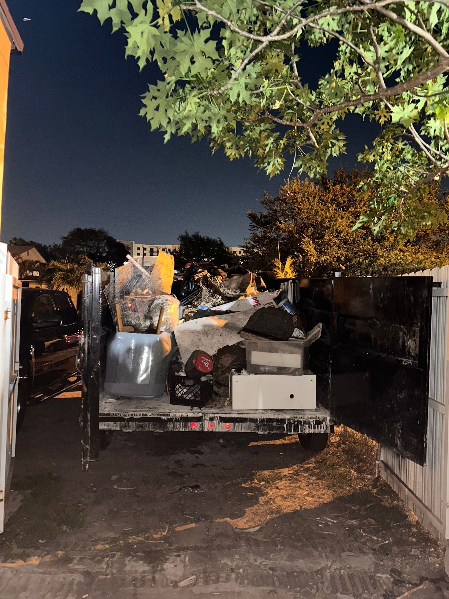 A black trailer filled with assorted household junk and debris parked in a driveway at night beneath tree branches.