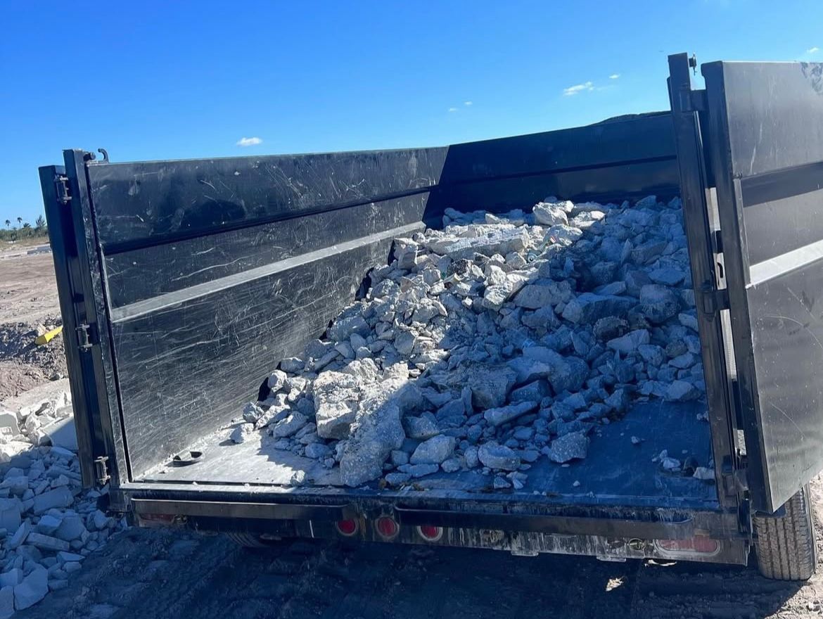 A black dump trailer loaded with jagged gray rocks parked on a sandy, sunlit construction site.