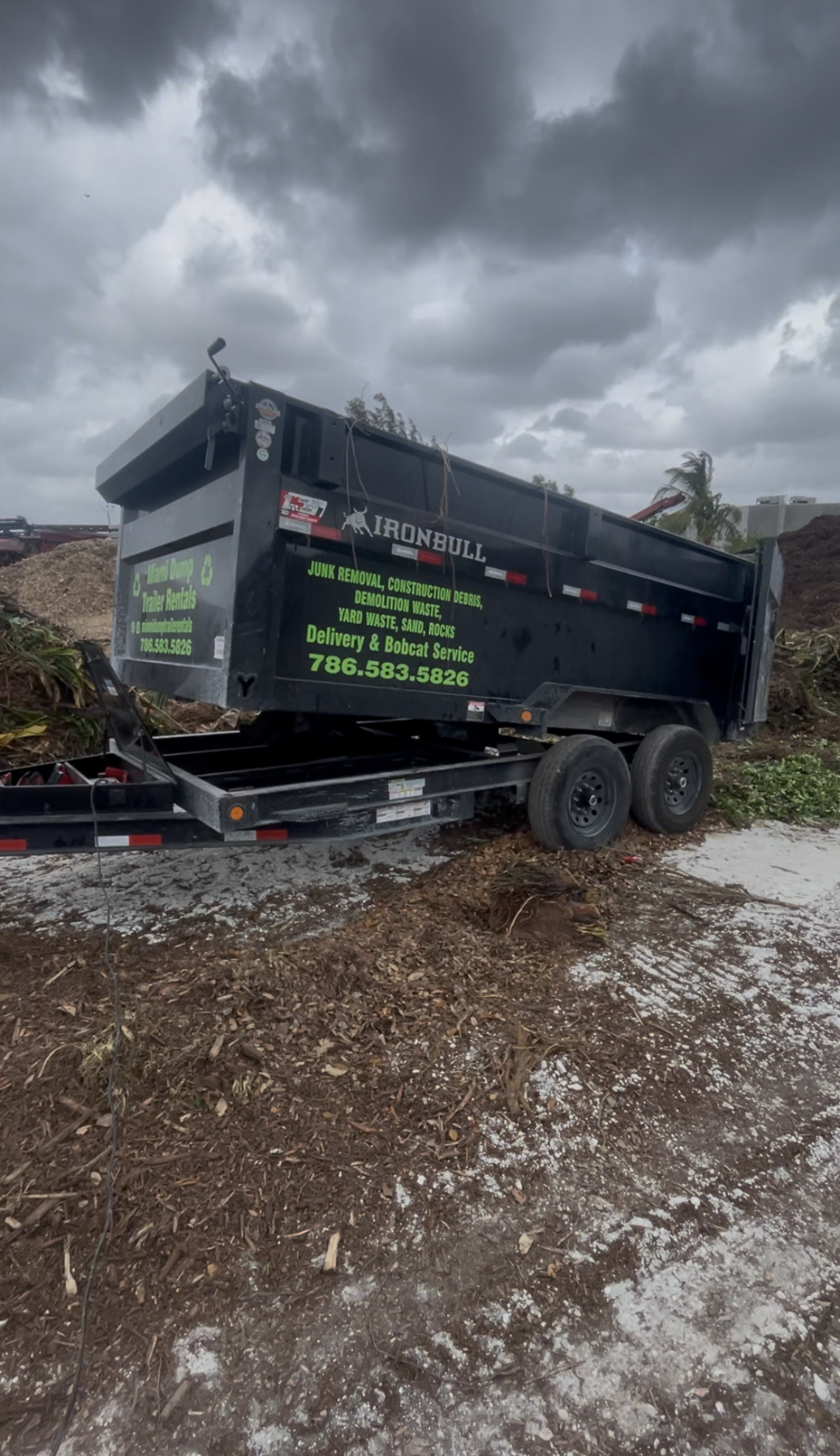 A black dump trailer parked on a dirt lot under a cloudy sky. The side displays company information and a phone number.